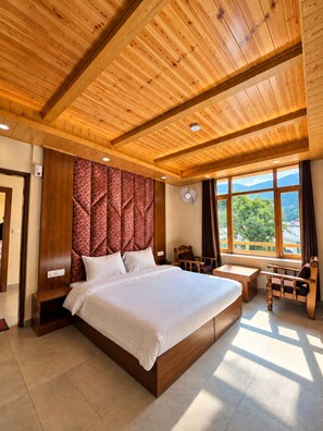 A spacious bedroom with a large bed, wooden ceiling beams, a window with a view of greenery, and a seating area with a table and chairs.