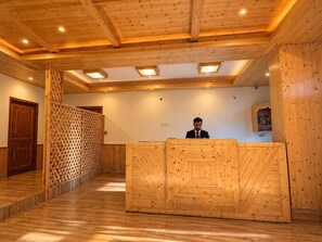Un mostrador de recepción de madera con una persona detrás, suelo de madera y techo con vigas de madera e iluminación empotrada.