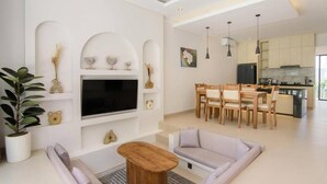 Una sala de estar moderna con un televisor de pantalla plana, una mesa de centro de madera y un sofá. Hay un comedor con sillas de madera y una cocina al fondo.