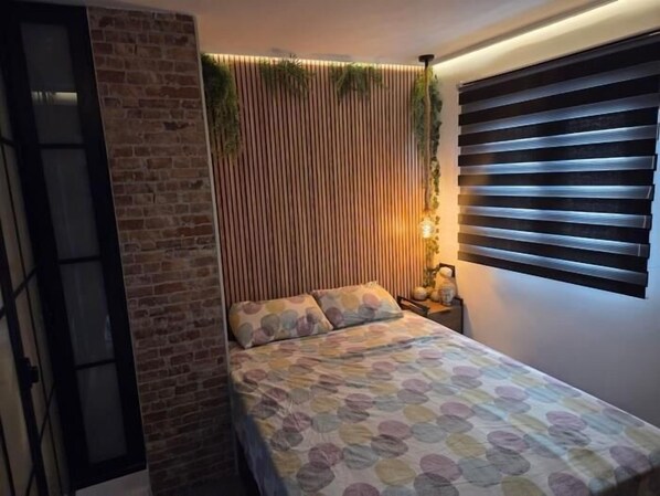 Une chambre à coucher avec un lit, une table de chevet, une plante fixée au mur et une fenêtre avec des stores.