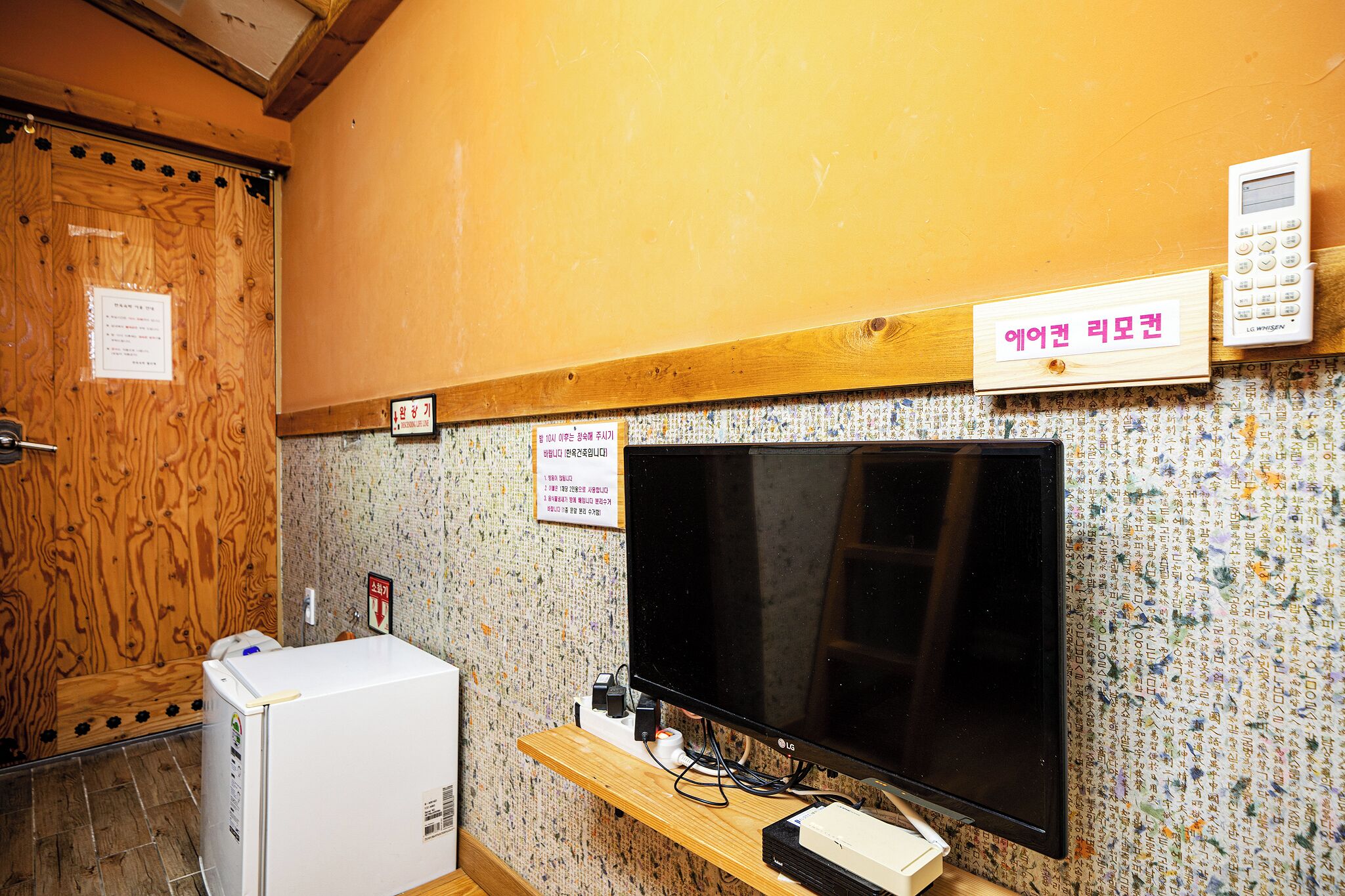 Ein Zimmer mit Fernseher, Kühlschrank und einem Holzkasten. An der Wand befinden sich Schilder und eine Tür mit einem Holzpaneel.