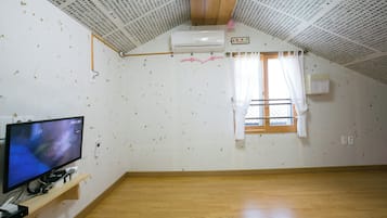 Een kamer met een schuine kap, een wandairconditioner, een raam met gordijnen en een televisie op een houten plank.