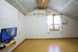 Una habitación con techo inclinado, aire acondicionado instalado, ventana con cortinas y televisor sobre un estante de madera.