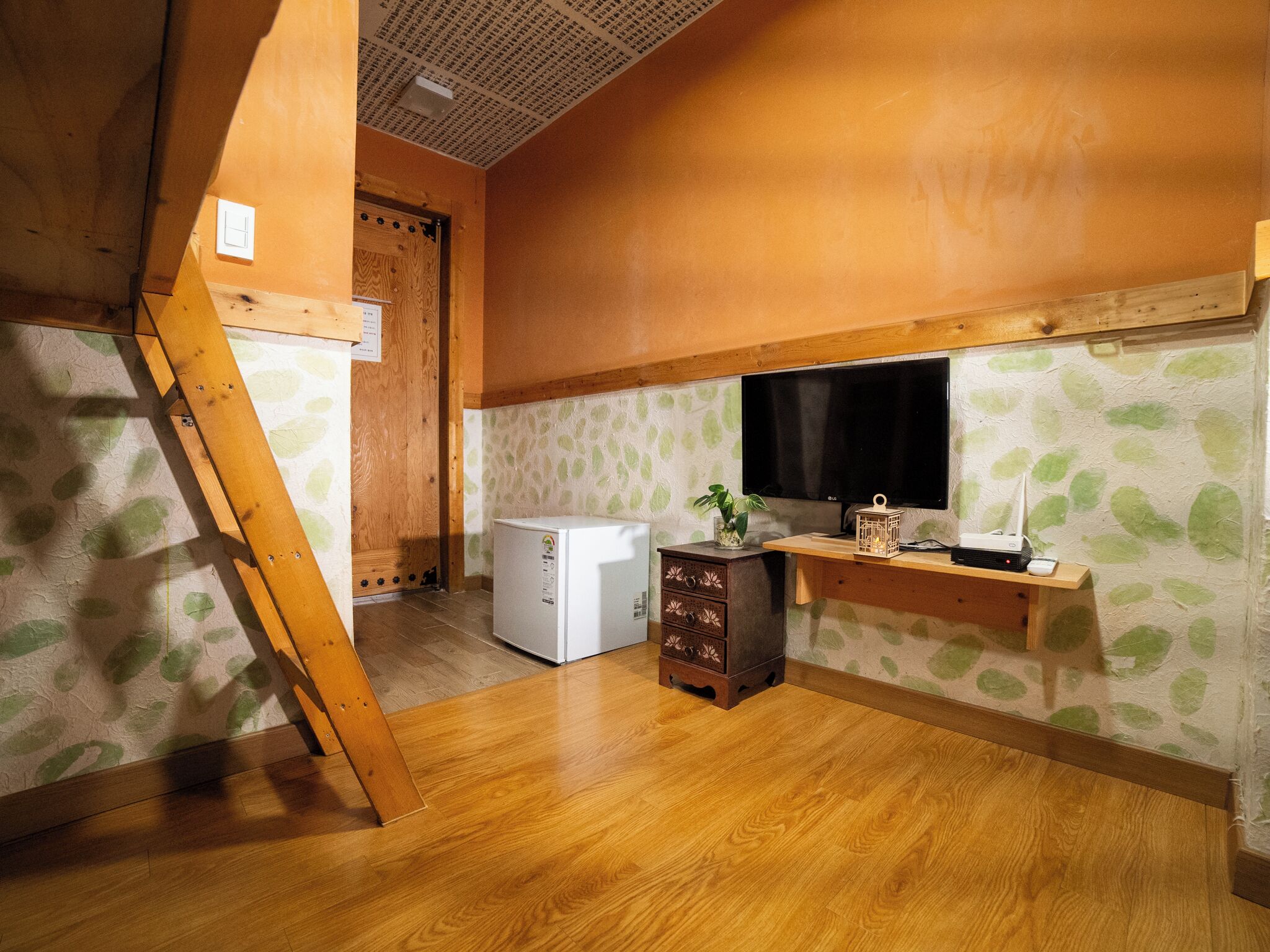 Una habitación con piso de madera, un refrigerador blanco, un gabinete de madera con cajones, un televisor sobre una repisa de madera y una puerta con marco de madera.