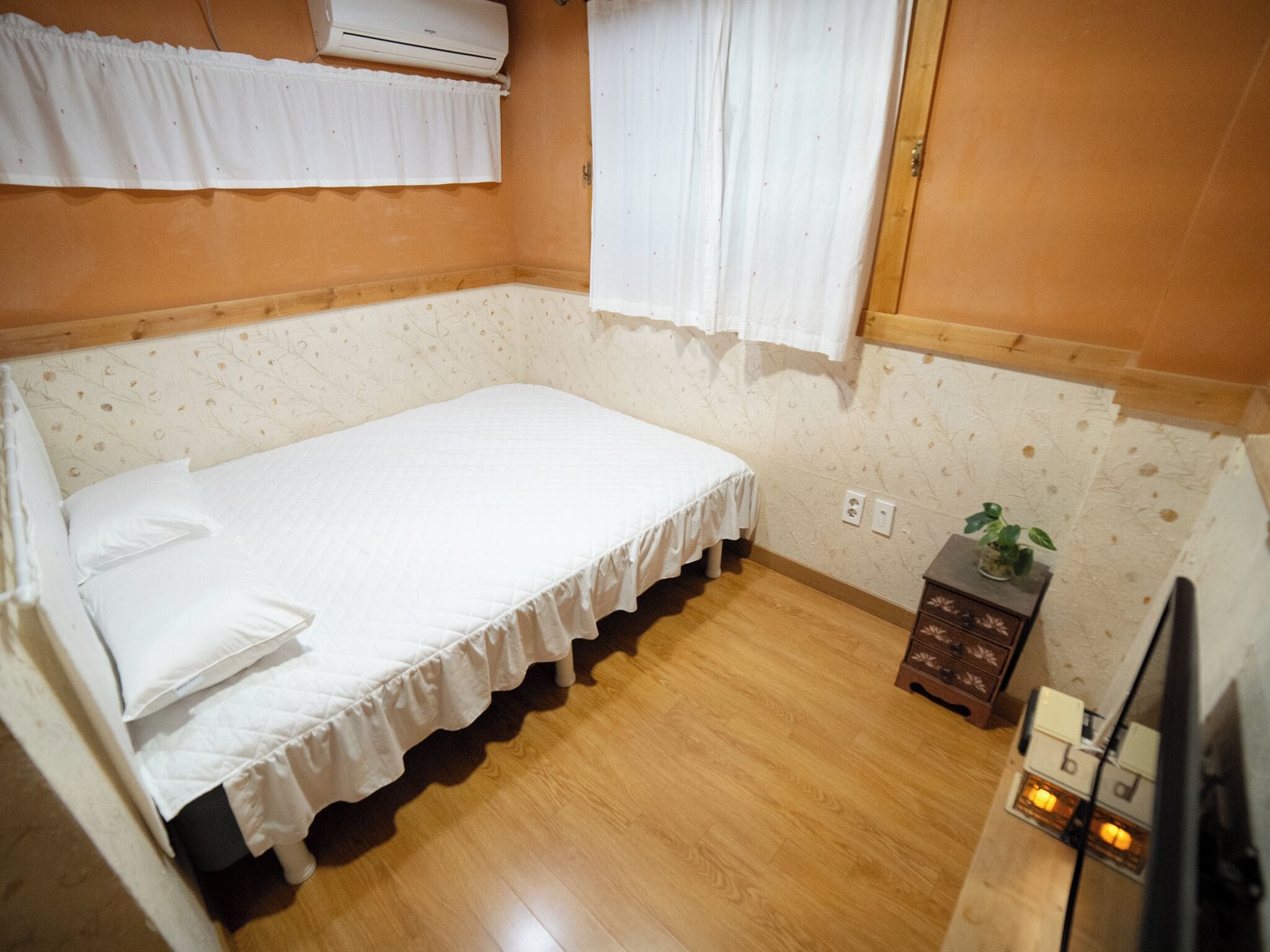 Una habitación pequeña con cama individual, piso de madera, edredón blanco y una mesita de noche con una planta.