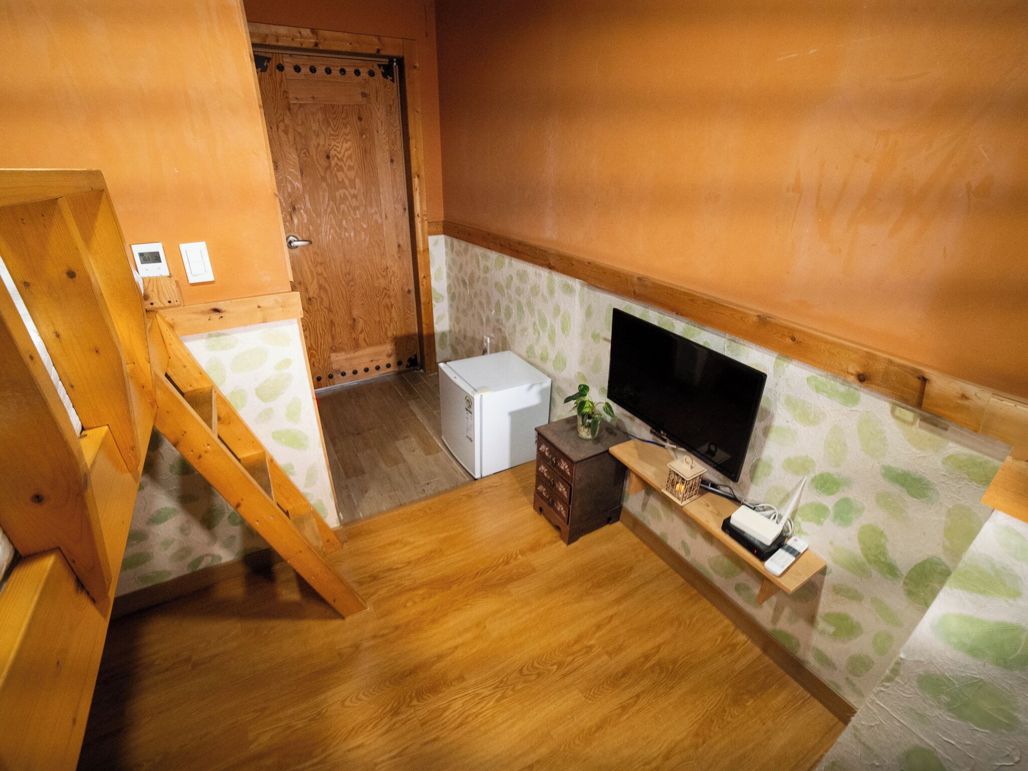 Habitación pequeña con suelo de madera, un refrigerador blanco y una televisión sobre una estantería, además de una puerta de madera.