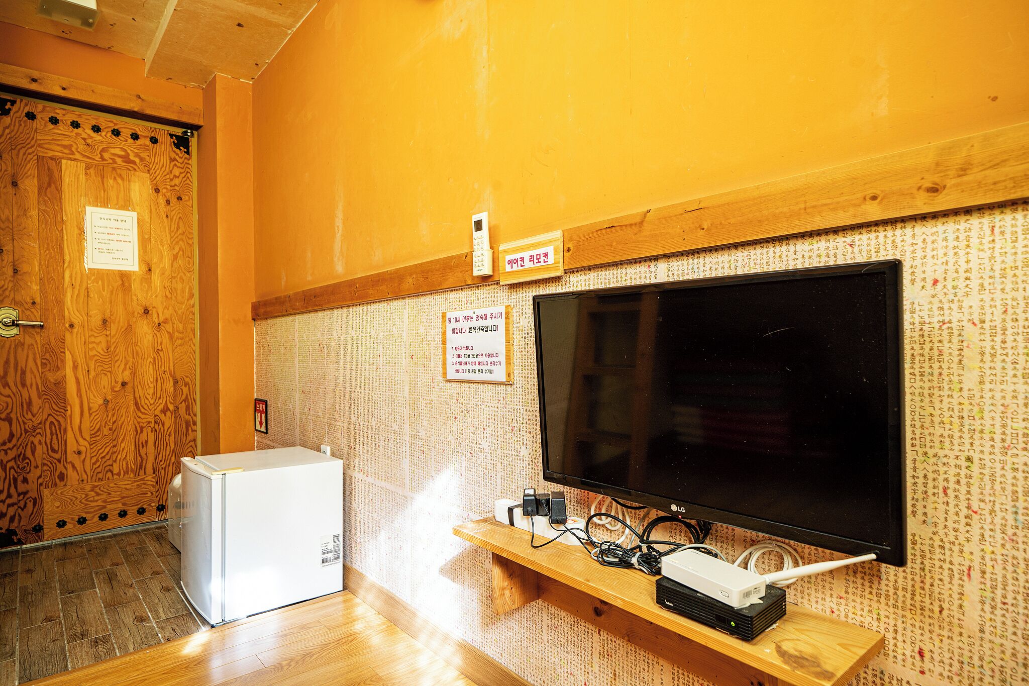 A room with a wooden door, a white refrigerator, a flat-screen TV on a wooden stand, and a sign with Korean text.