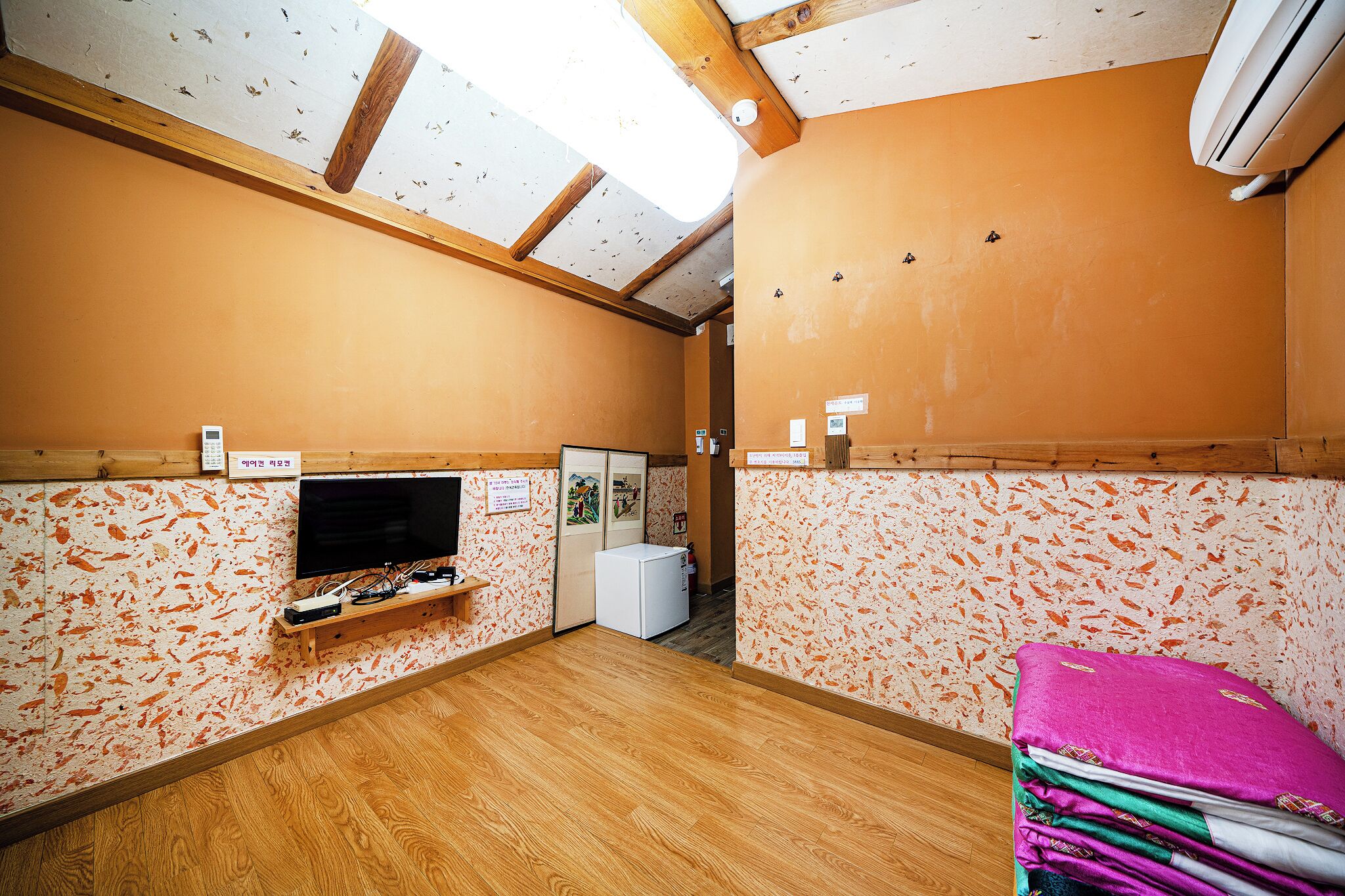 A room with a wooden ceiling, a television on a wooden shelf, a white refrigerator, and a stack of colorful towels.