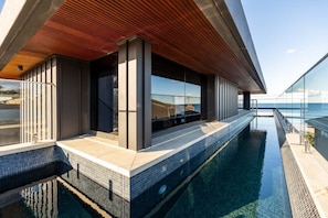 En modern infinitypool med ett träbeklätt tak och glasväggar, som erbjuder utsikt över havet.