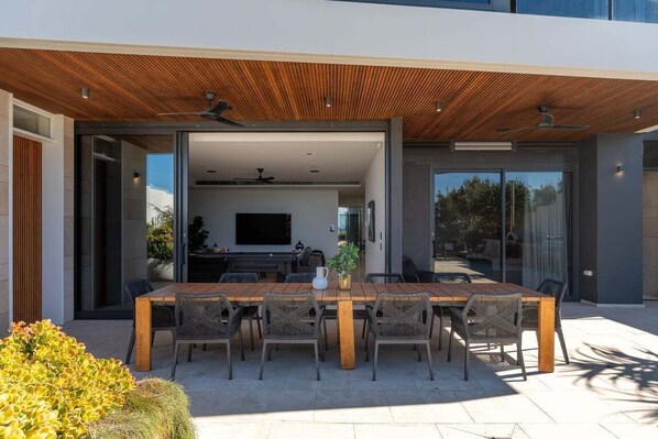 An outdoor dining area with a wooden table and chairs, a television in the background, and a view of the house's interior.