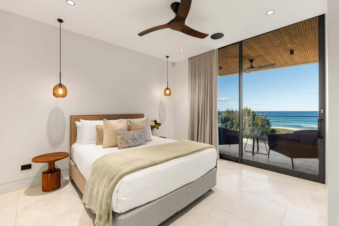 Une chambre moderne dotée d’un grand lit, d’un ventilateur de plafond et offrant une vue sur l’océan grâce à de grandes fenêtres du sol au plafond.