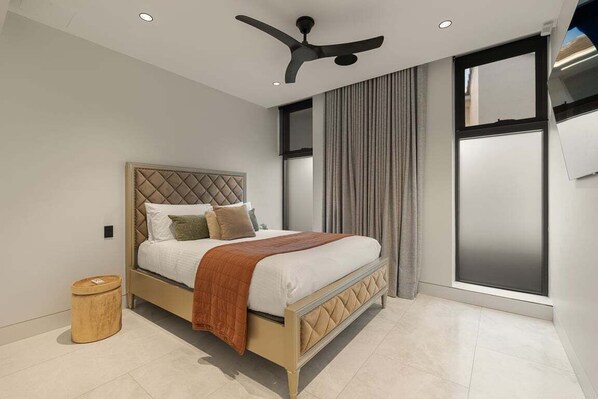 Un dormitorio con una cama, un ventilador de techo, una ventana con cortinas y un taburete de madera.