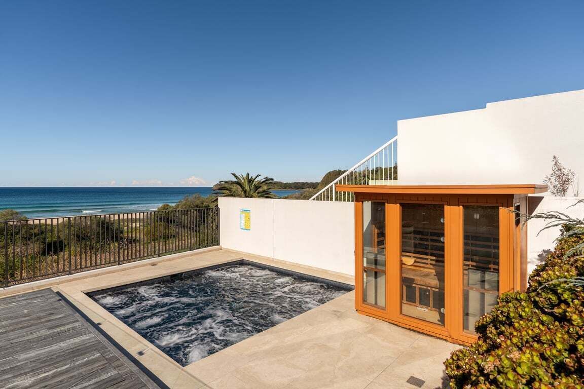 Un espace moderne aménagé à l’extérieur avec un jacuzzi, une vue sur la mer, une terrasse en bois et une structure fermée par des parois de verre.