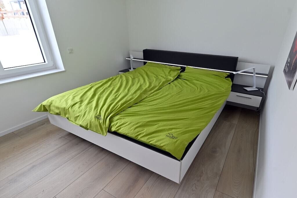 Un lit avec un cadre blanc et une couvre-lit verte, une table de chevet et une lampe fixée au mur.