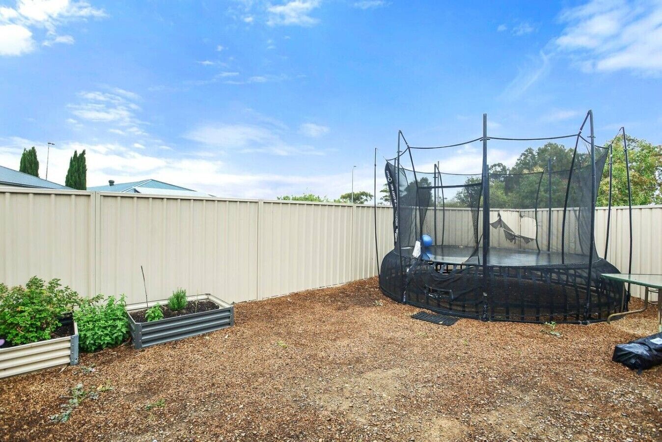 Un jardin à l’arrière de la maison, avec un trampoline entouré d’un filet de sécurité, délimité par une clôture en bois et agrémenté d’un parterre de fleurs.
