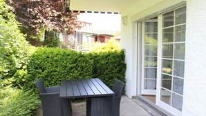 Eine Terrasse im Freien mit Tisch und Stühlen, umgeben von Grünpflanzen und einer weißen Gebäudefassade.
