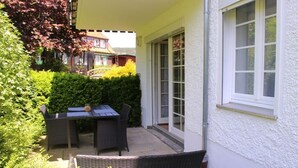 Eine Terrasse mit Tisch und Stühlen, eine weiße Außenwand und ein Heckenstreifen.