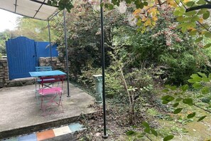 A covered patio area with a table and bench, surrounded by dense foliage and a stone wall. - Maffone la laupie gîte COCO (La Laupie)