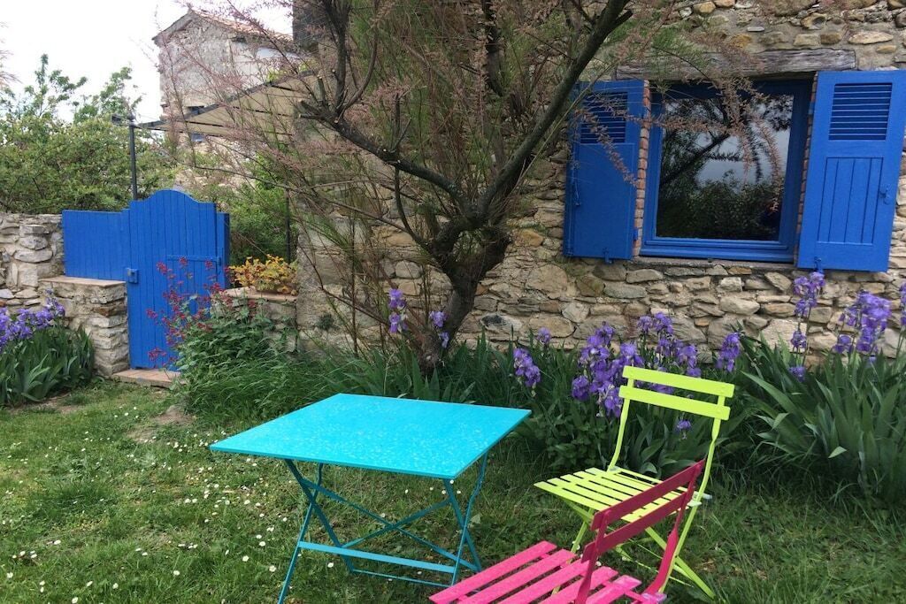 Un edificio de piedra con jardín, una reja azul y una mesa con una silla.