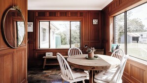 Uma área de refeições com uma mesa redonda de madeira, cadeiras brancas e uma janela grande com vista para o exterior.