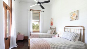 Quarto com duas camas, um ventilador de teto, janela com cortinas, mesa de cabeceira e um quadro pendurado na parede.