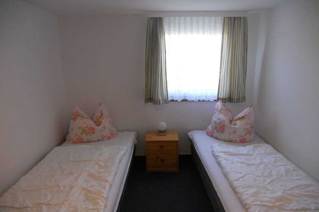 Een kleine, eenpersoonskamer met twee bedden, een nachtkastje en een raam met gordijnen.