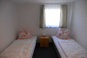 A small, single-room accommodation with two beds, a nightstand, and a window with curtains.