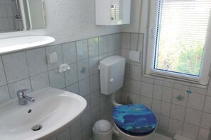 Ett badrum med ett vitt handfat, en toalett med ett blått sits och ett fönster med persienner.