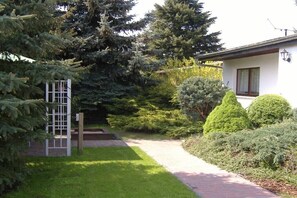 Una casa con camino de entrada pavimentado que lleva a una puerta blanca, rodeada de vegetación exuberante y árboles altos.