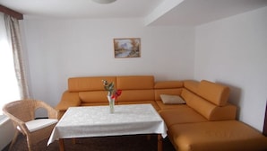 Um quarto com sofá seccional, uma mesinha com toalha branca, uma cadeira de vime e um quadro na parede.
