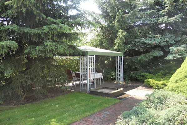 A covered outdoor seating area with a table and chairs, surrounded by dense trees.