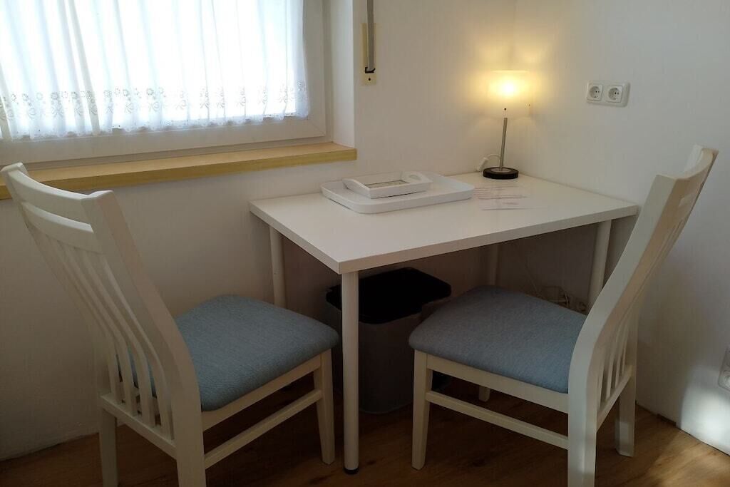 A small white table with a lamp, two chairs, a window with curtains, and a trash bin.