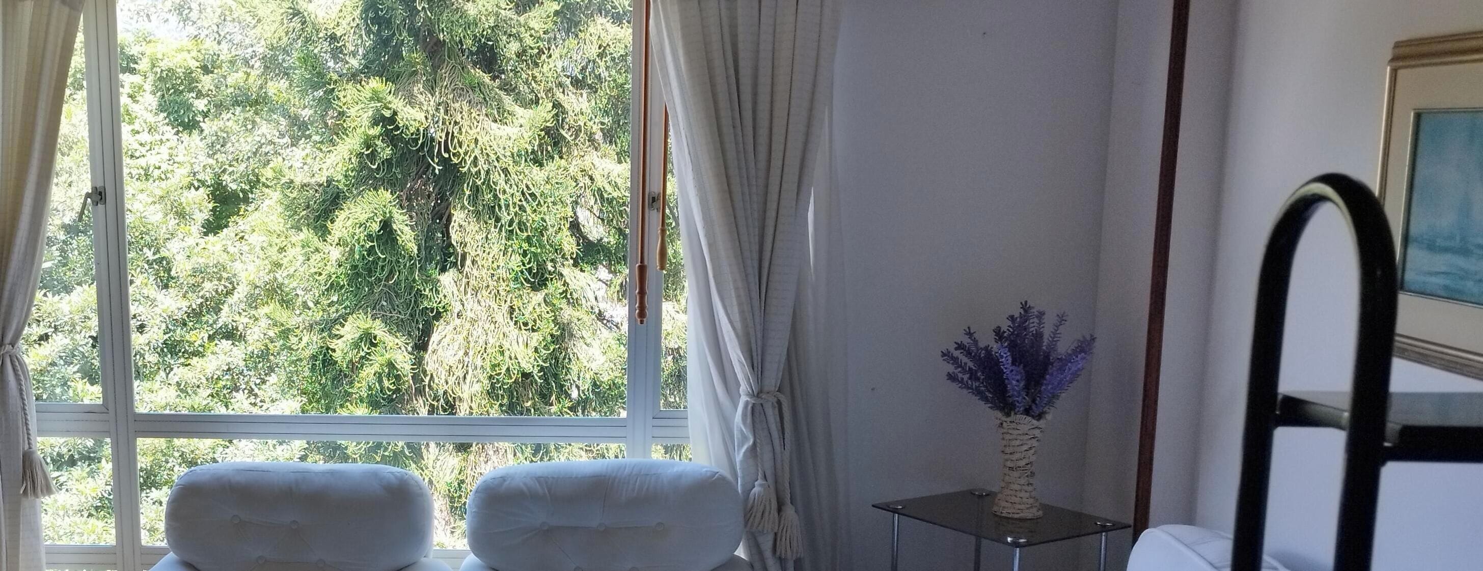 Una habitación con un gran ventanal, dos sillas blancas, una peque ña mesa con un florero y un espejo en la pared.