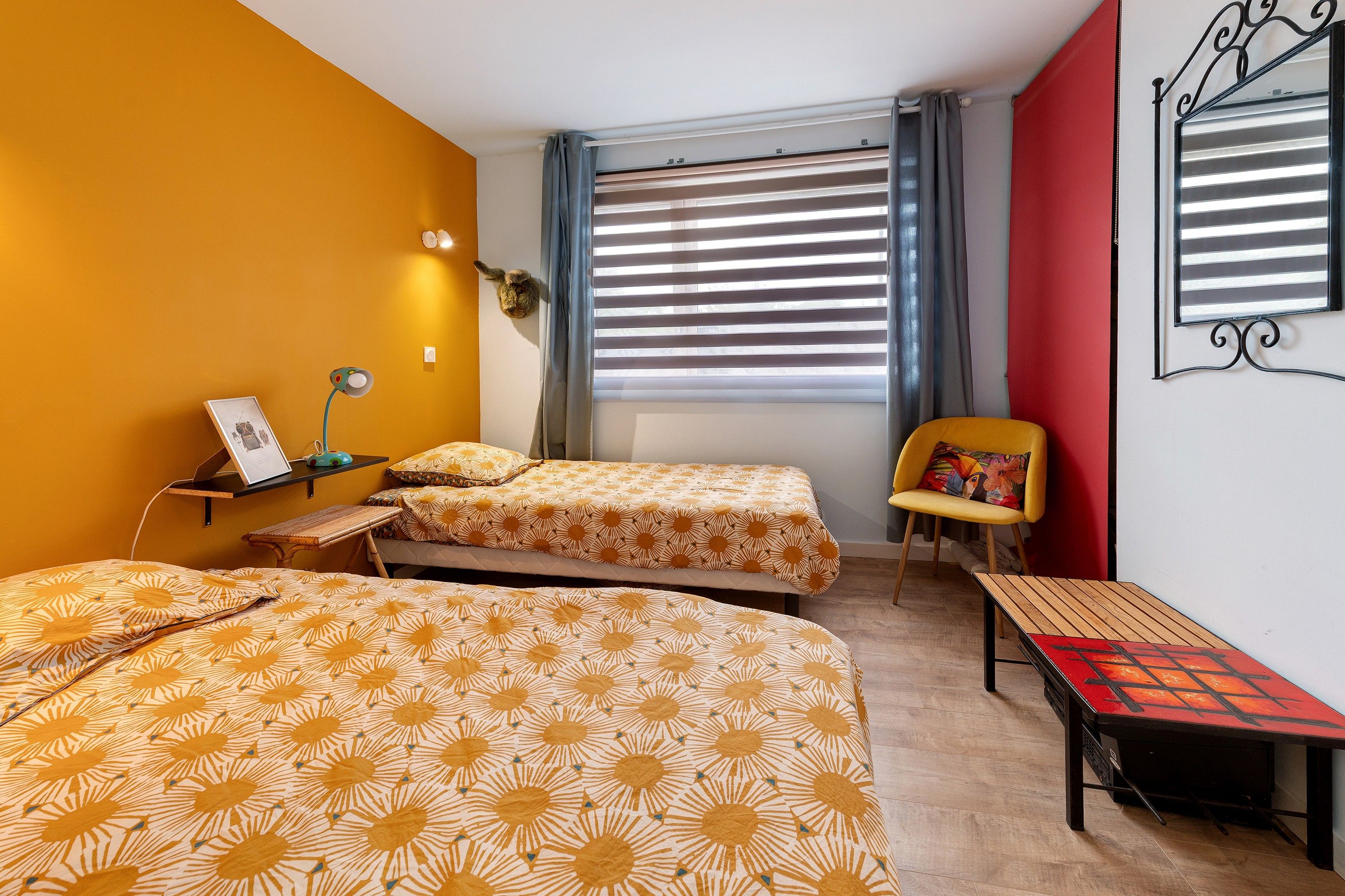 Ein Zimmer mit zwei Betten, einem Sessel, einem Tisch, einem Fenster mit Jalousien und einem wandmontierten Spiegel.