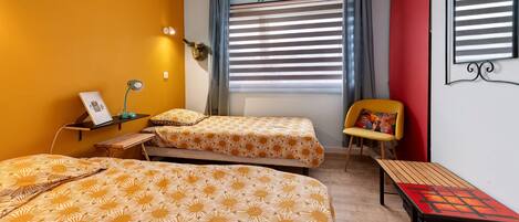 Ein Zimmer mit zwei Betten, einem Sessel, einem Tisch, einem Fenster mit Jalousien und einem wandmontierten Spiegel.