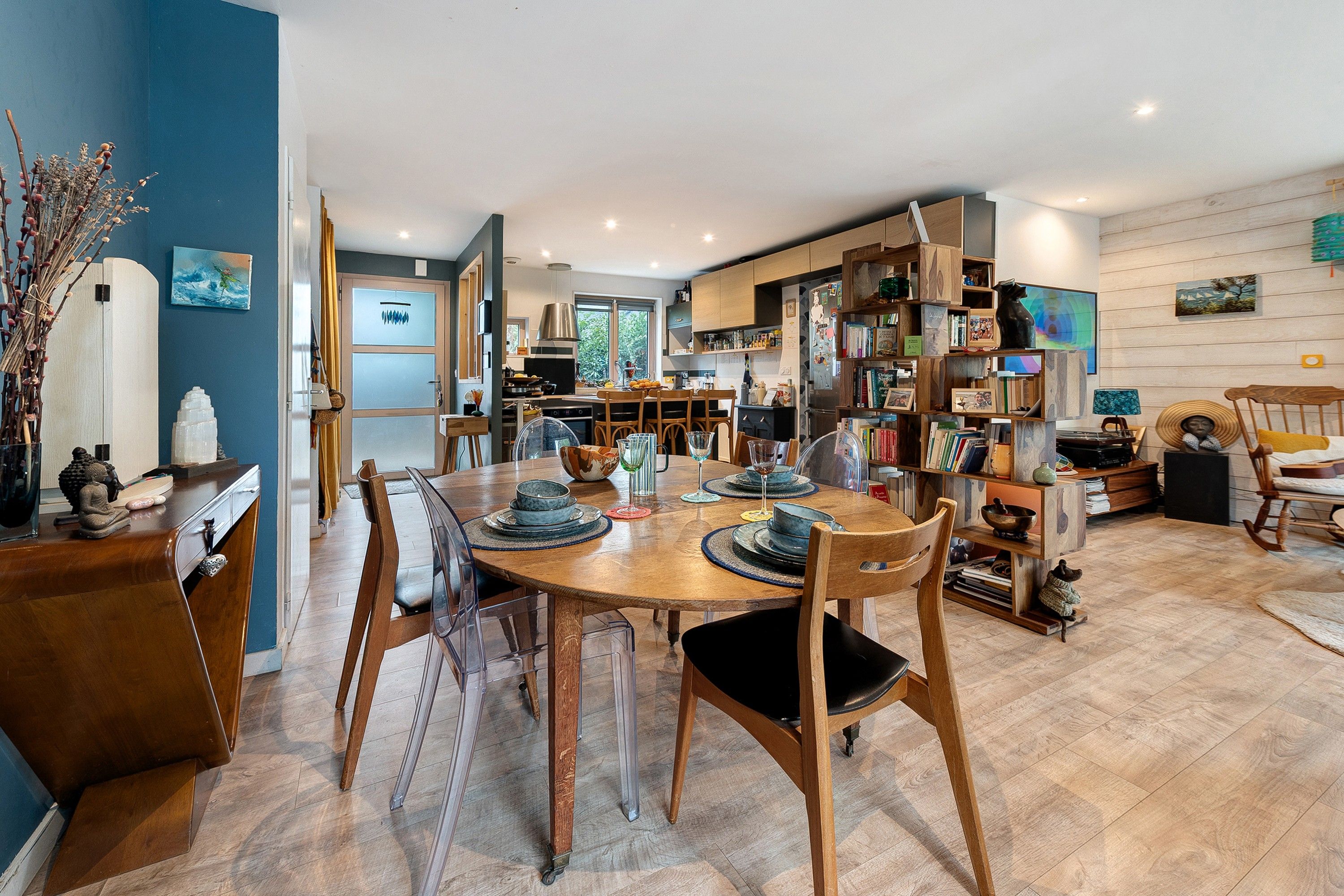 A modern kitchen with wooden cabinets, a dining area with a round table and chairs, and a living space with a bookshelf and decorative items.