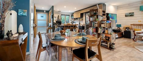 A modern kitchen with wooden cabinets, a dining area with a round table and chairs, and a living space with a bookshelf and decorative items.