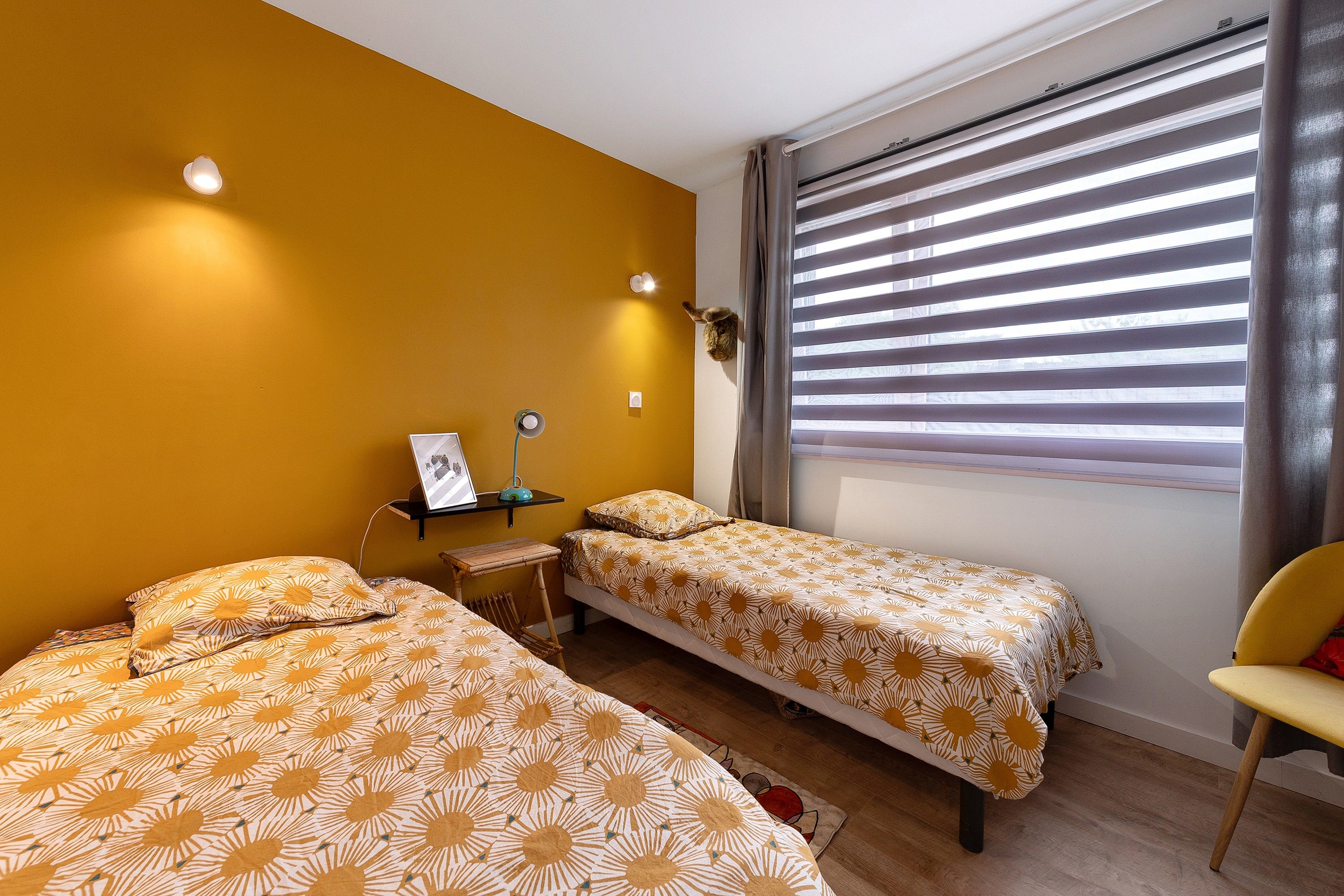 A room with two beds, a small table, a chair, and a window with blinds.