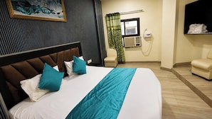 A hotel room with a large bed, a headboard, a window with an air conditioning unit, a green curtain, a wall-mounted TV, and a seating area with a sofa and chairs. - HOTE NATRAJ INN  (Varanasi)