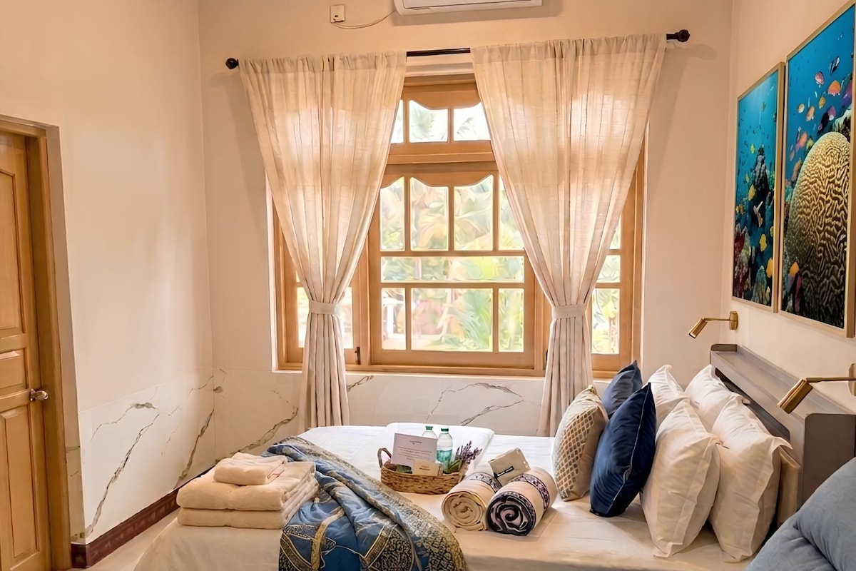 A hotel room with a bed, a sofa, a window with curtains, an air conditioning unit, and artwork on the wall.