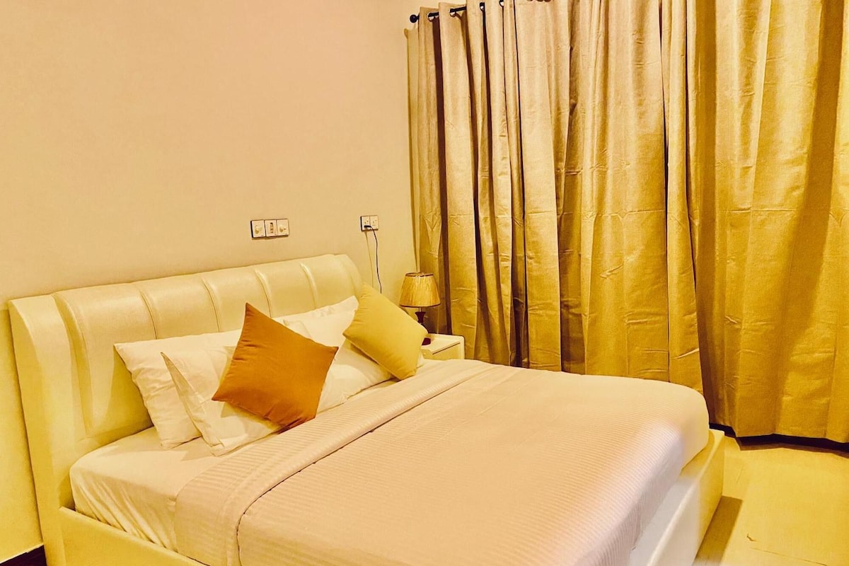 A hotel room with a bed, pillows, a nightstand, a lamp, and curtains.