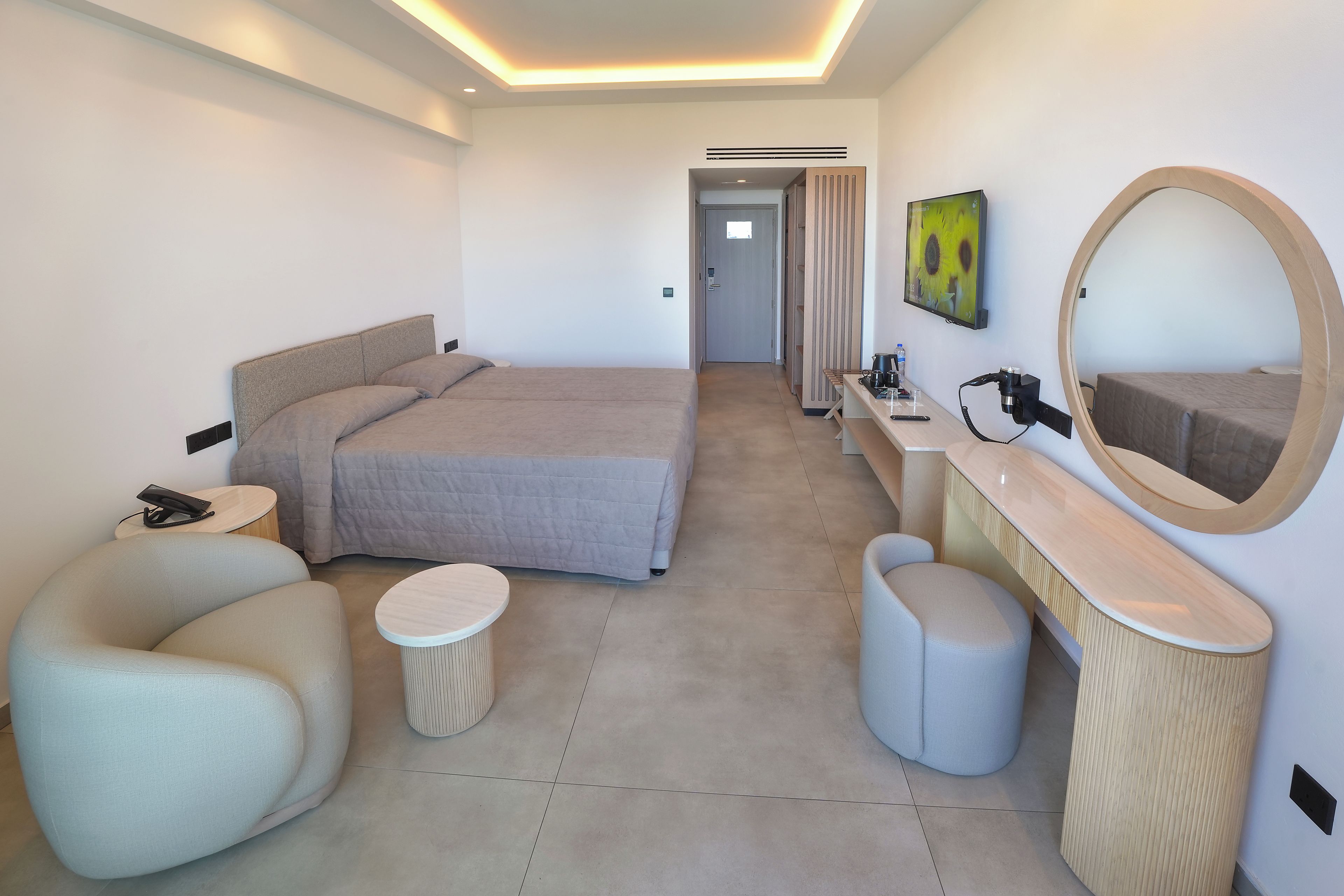 A modern hotel room with a bed, a desk, a chair, a small round table, a mirror, and a painting on the wall.