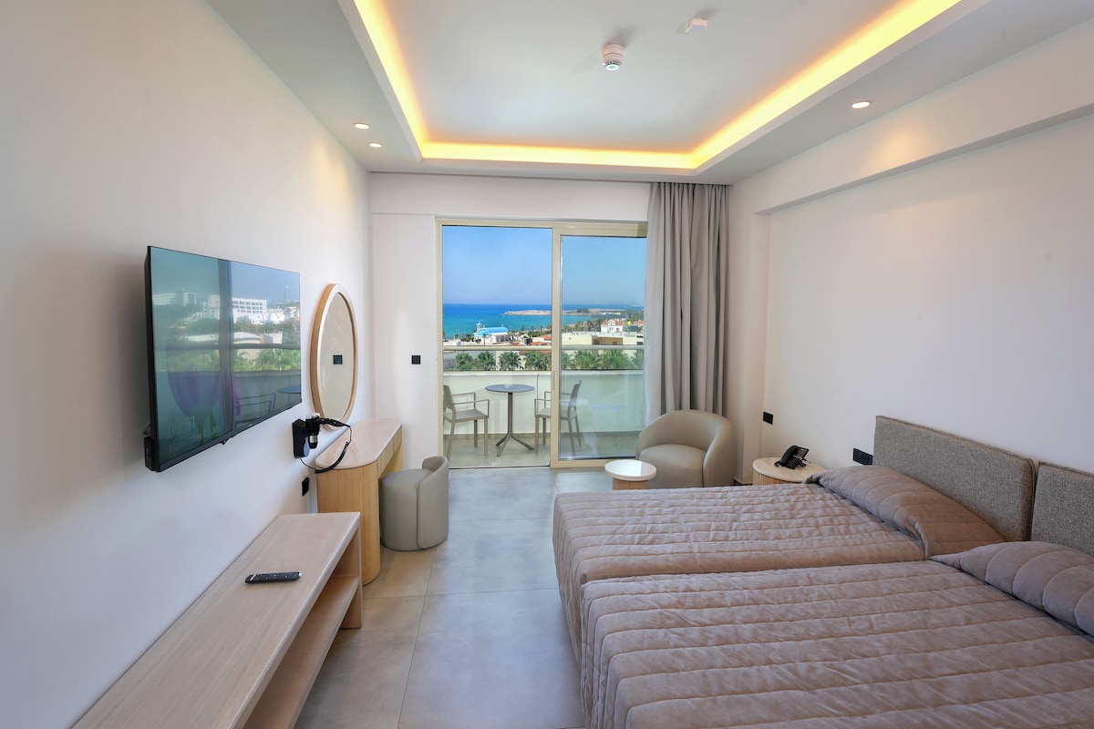 Quarto de hotel com cama, escrivaninha, cadeira, televisão e varanda com vista para o mar.
