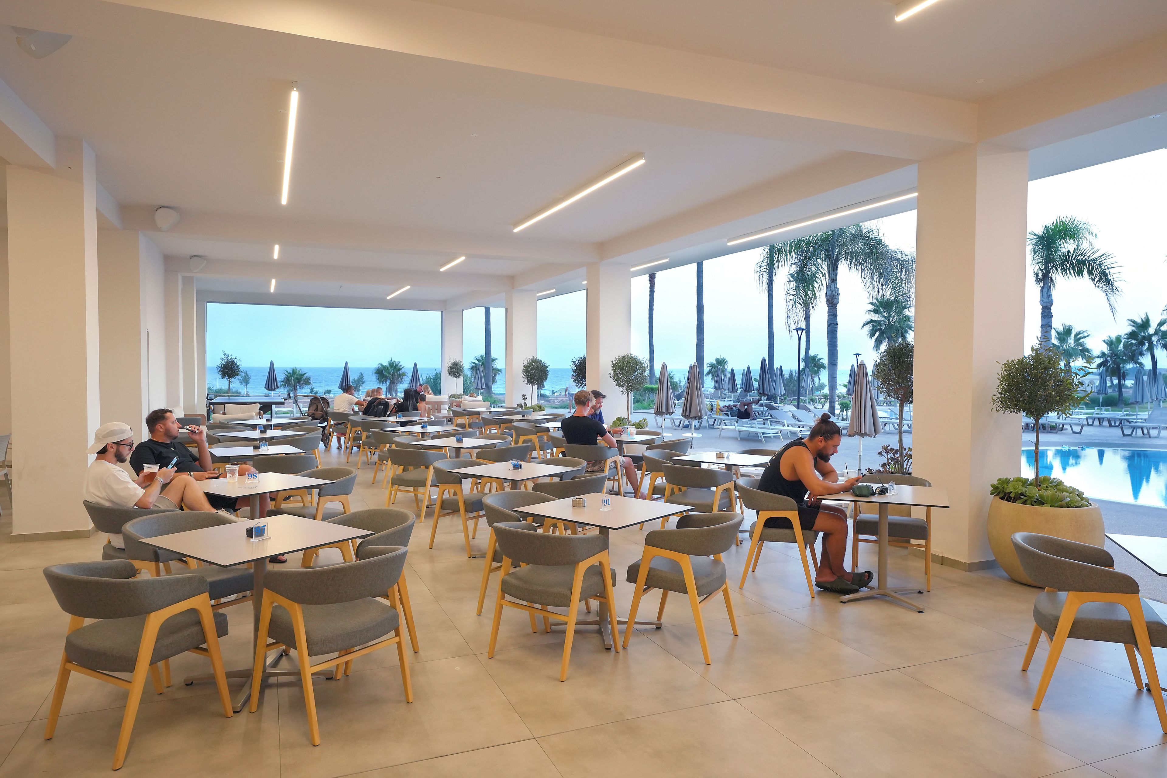 Un amplio comedor interior con múltiples mesas y sillas, ventanas grandes que ofrecen vista al mar, y un área junto a la piscina con sombrillas.