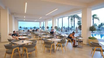 Un amplio comedor interior con múltiples mesas y sillas, ventanas grandes que ofrecen vista al mar, y un área junto a la piscina con sombrillas.