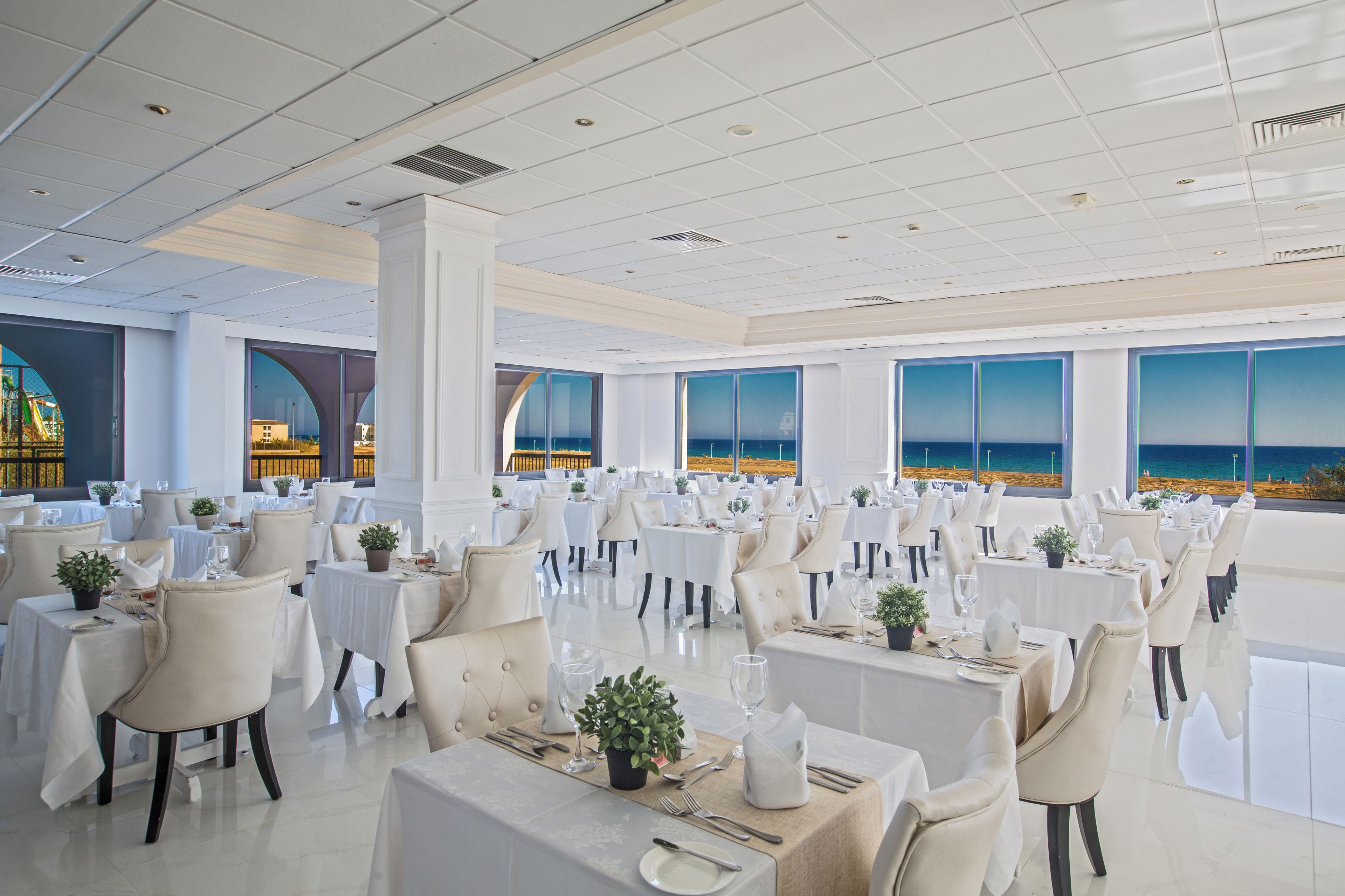 A spacious dining area with tables set for service, large windows offering a view of the sea, and a modern ceiling design.
