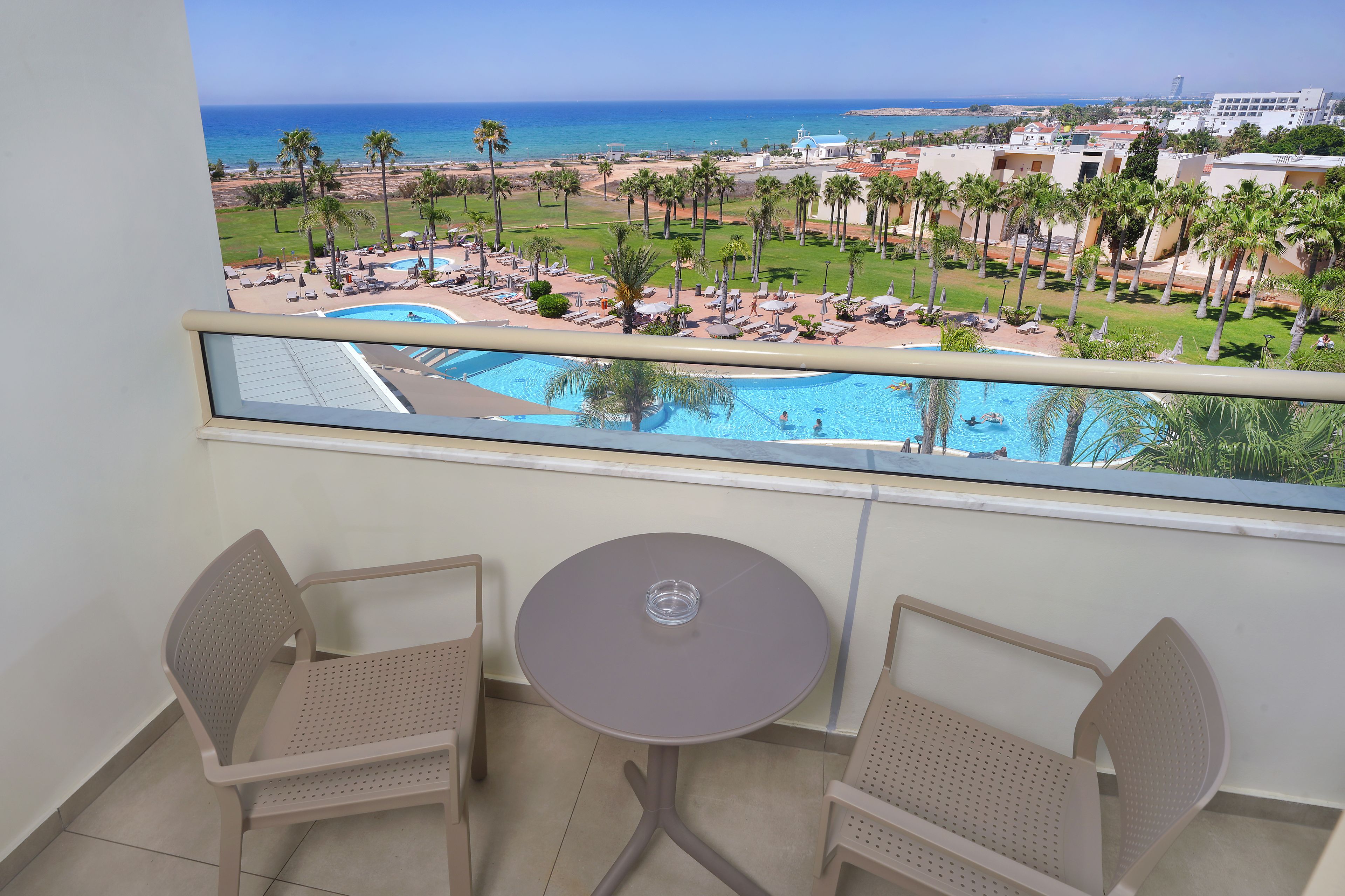 Un balcón con dos sillas y una mesita, con vistas a una zona de piscina con palmeras y el mar.