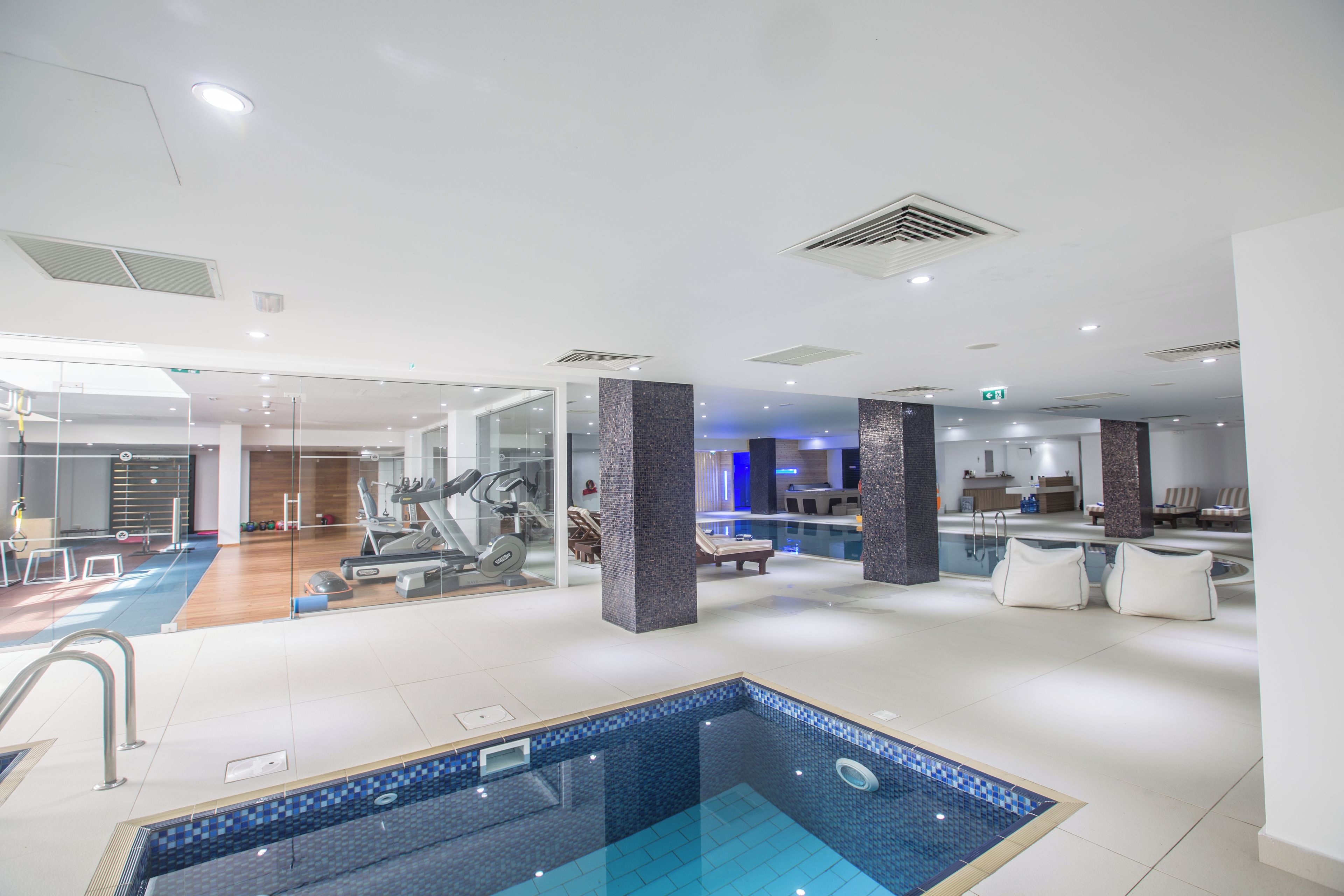 Una piscina interior moderna con cerramiento de vidrio, rodeada de equipo de ejercicio y zonas de descanso.