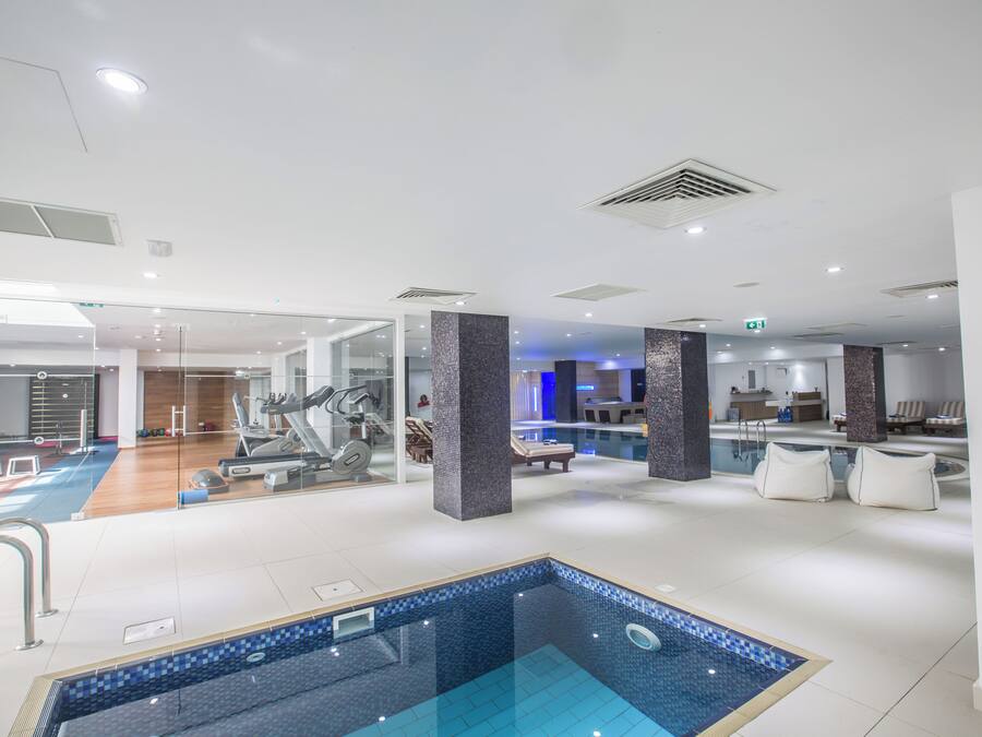 Una piscina interior moderna con cerramiento de vidrio, rodeada de equipo de ejercicio y zonas de descanso.