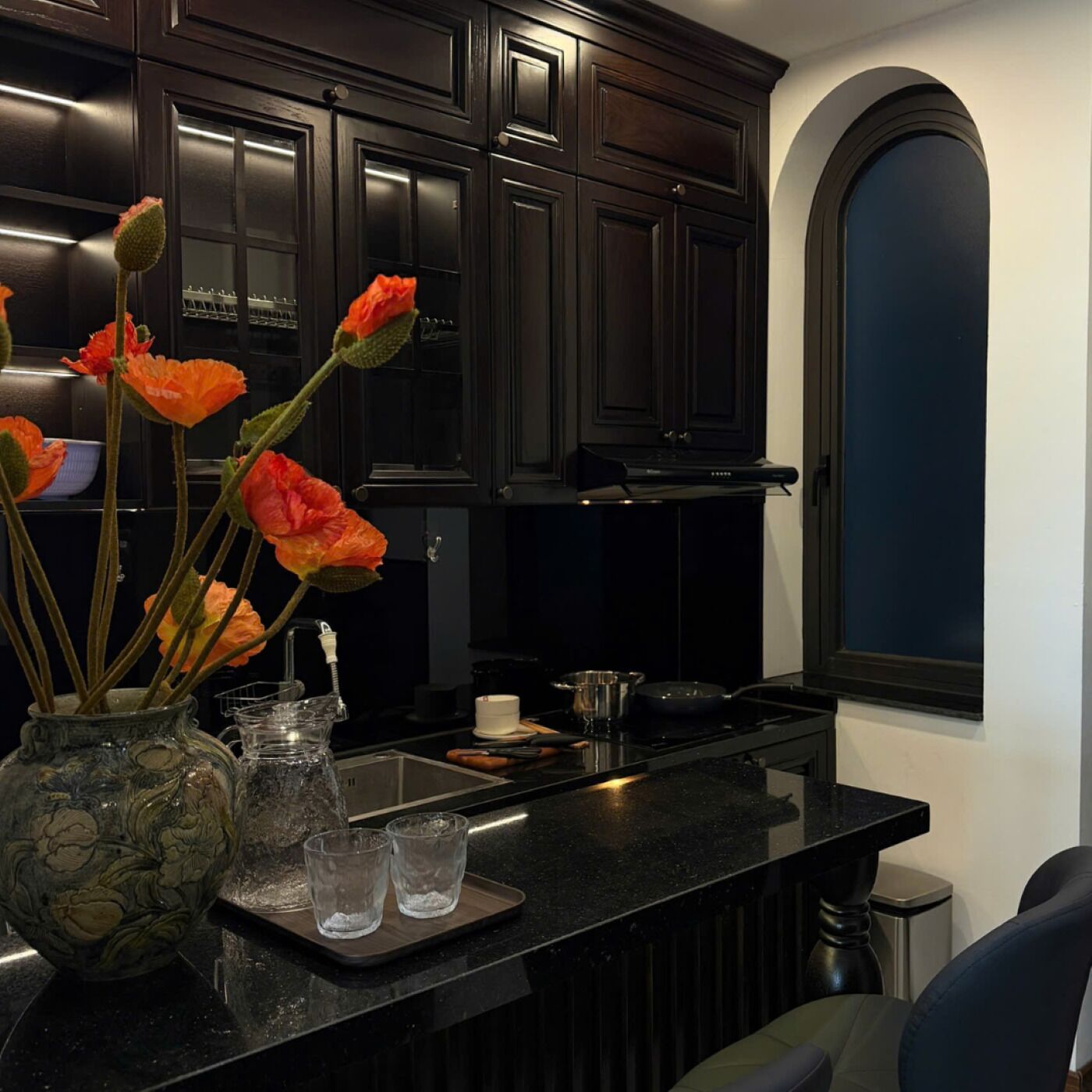 A kitchen with dark wooden cabinets, a black countertop, a vase with orange flowers, and a clear glass pitcher.